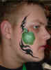 Face Painting: Adult male with cheek painted green apple framed in black trible pattern.